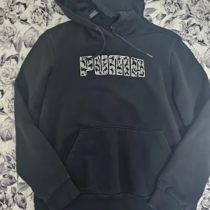Svart hoodie från Puma med leopardmönster - Svart hoodie från Puma i storlek M. Tröjan har huva med dragsko, magficka och ett stort PUMA-tryck i vitt med leopardmönster på bröstet. Perfekt för en chill och sportig stil.