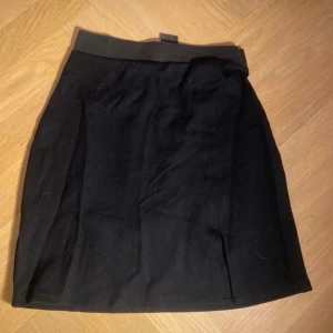 Svart kortkjol med slits - Enkel svart kortkjol med diskret slits framtill och bred midjeresår. Kjolen har en rak passform och är tillverkad i ett mjukt tyg som känns bekvämt mot huden. Perfekt att matcha med en stickad tröja eller t-shirt.