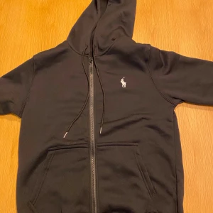 Svart hoodie och svart byxa  från Polo Ralph Lauren - Svart hoodie från Polo Ralph Lauren med klassisk logga broderad i vitt på bröstet. Tröjan har dragkedja framtill, huva och fickor. Materialet är mjuk bomull som känns skönt mot huden. Perfekt för dig som gillar stilrena och tidlösa plagg.