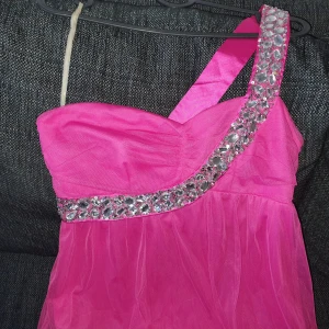 Rosa långklänning med strass från Bodyflirt - Snygg rosa långklänning från Bodyflirt med ett axelband dekorerat med stora strassdetaljer. Klänningen har oneshoulder-modell och är sydd i ett tunt, lätt tyg som ger ett flowy intryck. Perfekt för dig som vill sticka ut med färg och glitter.