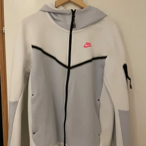 Nike hoodie med dragkedja och huva - Nike hoodie i vitt och ljusgrått med svart dragkedja och huva. Tröjan har lång ärm, fickor och en rosa Nike-logga på bröstet. Snygga svarta detaljer och en ficka med dragkedja på ena ärmen.