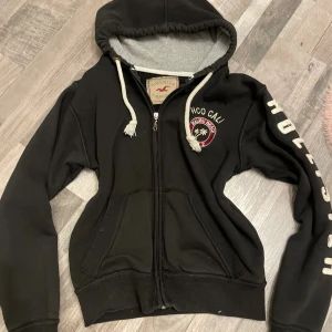 Svart hoodie Hollister Malibu Beach - Svart hoodie från Hollister med broderad Malibu Beach-logga på bröstet och stora vita bokstäver på ena ärmen. Tröjan har huva med vita snören, dragkedja framtill och två fickor. Insidan är mjuk och grå, perfekt för chill dagar.