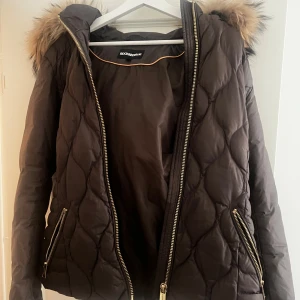  pufferjacka Rockandblue - Snygg brun pufferjacka från Rockandblue med quiltad design och fluffig fuskpäls på huvan. Jackan har guldfärgade dragkedjor, två fickor fram och en badge på ärmen. Perfekt för kalla dagar med sin varma och stilrena look.