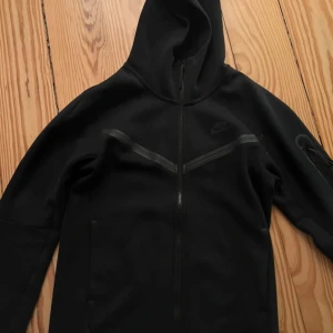 Svart hoodie från Nike med dragkedja - Svart hoodie från Nike med hel dragkedja och huva. Snygga detaljer med svarta linjer över bröstet och en ficka med dragkedja på ärmen. Långärmad och tillverkad i mjukt material som känns skönt mot huden. Perfekt för chill eller träning.