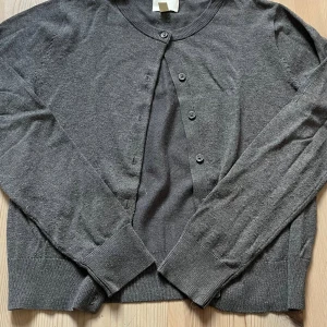 Mörkgrå kofta med knappar - En stilren mörkgrå kofta med rund hals och långa ärmar. Koftan har ribbade muddar och knäppning med fyra knappar framtill. Perfekt att slänga över en t-shirt eller linne för en chill look.