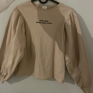 Beige långärmad tröja från Zara - Beige långärmad tröja från Zara med puffiga ärmar och svart broderad text på bröstet: 'Great plans become great stories.' Rund halsringning och avslappnad passform, perfekt för en chill och stilren look.