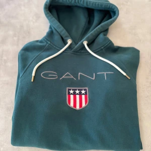 Mörkgrön hoodie från GANT med tryck - Snygg mörkgrön hoodie från GANT med broderad logga och emblem på bröstet. Tröjan har huva med vita snören och en stor magficka framtill. Tillverkad i mjuk bomull, perfekt för chill dagar. Klassisk och stilren design med coola detaljer. Nypris 900kr