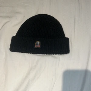 Svart ribbad mössa Parajumpers - Svart ribbstickad mössa från Parajumpers med uppvikt kant och klassisk PJS-logga framtill. Tillverkad i mjukt material som håller dig varm och snygg. Perfekt accessoar för kyliga dagar och enkel att matcha med din jacka.