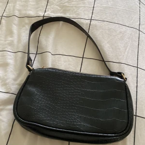 Svart axelväska i croco-look från H&M - Svart axelväska från H&M med croco-mönstrad yta och guldfärgad dragkedja. Väskan har ett hjärtformat hänge vid dragkedjan och är tillverkad i syntetmaterial. Perfekt storlek för det viktigaste och snygg detalj till din outfit. 