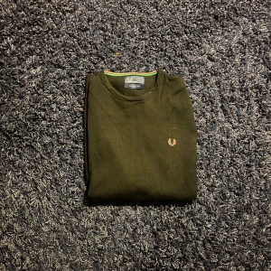Svart merinoulltröja Fred Perry XL - Svart tröja från Fred Perry i 100% merinoull, med klassisk rund hals och diskret logga broderad på bröstet. Halsringningen har en snygg detalj med röd och grön rand. Perfekt för dig som gillar stilrena och tidlösa plagg.