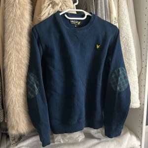 Blå sweatshirt från Lyle & Scott - Mörkblå sweatshirt från Lyle & Scott med broderad gul logga på bröstet och rutiga armbågslappar i grönt och rött. Tröjan har rund hals och långa ärmar, perfekt för en chill och stilren look.