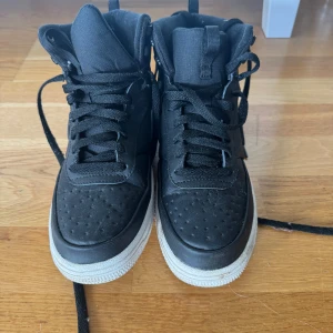 Nike Air Force 1 Mid svart/vit - Säljer ett par svarta Nike Air Force 1 Mid med vit sula. Perfekta till vintern. Skorna har klassisk snörning, rund tå och är tillverkade i en mix av skinn och syntet. Swoosh-logga på sidan och Nike-detaljer på sulan. Perfekta för dig som gillar streetstyle och vill ha en ikonisk sneaker. Ganska oanvända och i väldigt bra skick, helt onött sula. Priset är diskuterbart