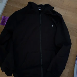 Svart hoodie från Polo Ralph Lauren - Svart hoodie från Polo Ralph Lauren med dragkedja framtill och klassisk vit broderad logga på bröstet. Tröjan har huva, känguruficka och är tillverkad i mjuk bomullsmix. Perfekt för en chill och stilren look.