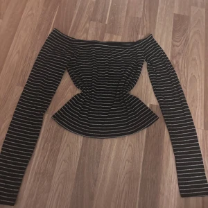 Randig offshoulder topp Noisy May - Säljer en svart offshoulder topp från Noisy May med vita horisontella ränder. Toppen har lång ärm och en figurnära passform som markerar midjan. Perfekt för dig som gillar en trendig och stilren look.