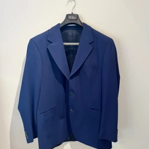 Blå kavaj från Tiger of Sweden - Snygg marinblå kavaj från Tiger of Sweden med klassisk passform och två knappar framtill. Kavajen har smala slag, tre fickor och är tillverkad i ett slätt material med diskret struktur. Perfekt för dig som gillar stilrena och tidlösa plagg. Det är en gammal kavaj och har tvärr två små hål i en ärm annars perfekt skick. Hör av er vid frågor 