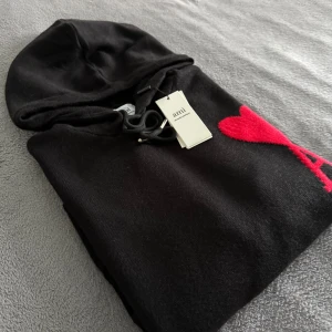 Ami Paris tröja  - Svart hoodie från Ami Alexandre Mattiussi med stor röd hjärtapplikation och bokstaven A på bröstet. Tröjan har huva med snörning, ribbade muddar och en klassisk känguruficka framtill. Materialet är mjukt och stickat, perfekt för chill dagar.