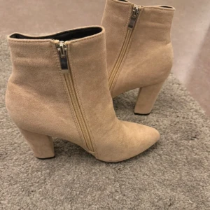 Beiga mocka stövletter med klack - Snygga beiga stövletter i mocka med spetsig tå och hög klack. Skorna har dragkedja på sidan för enkel på- och avtagning. Perfekta för att lyfta din outfit med en stilren och trendig look.