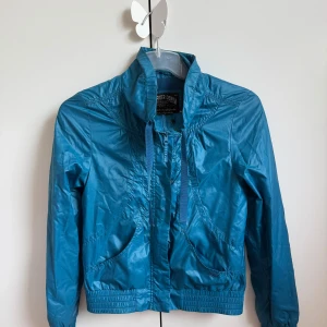 Blå vindjacka från Hilfiger Denim - Snygg blå vindjacka från Hilfiger Denim med hög krage, dragsko och dragkedja framtill. Jackan har elastiska muddar vid ärmslut och nederkant, samt två framfickor. Materialet är glansigt och lätt, perfekt för blåsiga dagar.