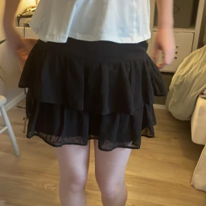 Svart volangkjol från Gina Tricot - Supersöt svart kortkjol från Gina Tricot med flera lager volanger och diskret transparent nederkant. Kjolen har dragkedja i sidan och är gjord i ett lätt, flowy material som ger en snygg siluett. Perfekt att styla med en enkel topp eller stickad tröja.