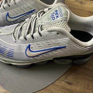 Nike Air Vapormax 360 vita/blå sneakers - Nike Air Vapormax 360 sneakers i vitt med blå detaljer och silverfärgad mesh. Skorna har en futuristisk design med genomskinlig, bubblig sula och blå Swoosh-logga. Ovandelen är i syntet och mesh med snörning och coola linjemönster.