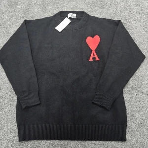 Svart stickad tröja från Ami Paris - Svart stickad tröja från Ami Paris med röd hjärtlogga och bokstaven A på bröstet. Rund halsringning och ribbade muddar vid ärmslut och nederkant. Perfekt för dig som gillar stilrena plagg med en twist.