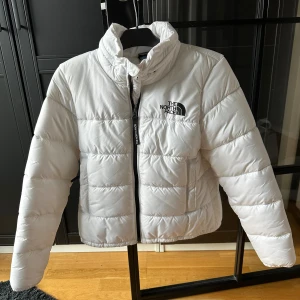 Vit pufferjacka från The North Face - Säljer en snygg vit pufferjacka från The North Face med hög krage och svart logga på bröstet. Jackan har dragkedja framtill, två stora fickor och quiltad design. Perfekt för kalla dagar och har en clean, modern look.