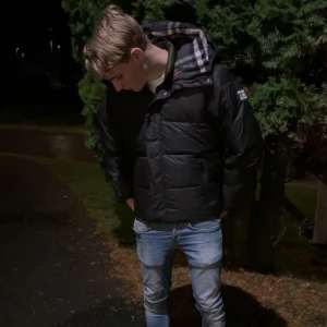 Svart pufferjacka från Burberry - Säljer en svart pufferjacka från Burberry med klassiskt rutigt foder i huvan. Jackan har dragkedja och tryckknappar framtill samt en tydlig Burberry-patch på ärmen. Perfekt för kalla dagar och riktigt snygg streetstil. Kan även göras till väst.