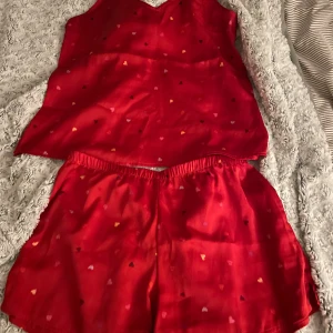 Röd hjärtmönstrad pyjamas - Säljer en pyjamas med linne o shorts i storlek XS men passar även större. De har ett gulligt hjärtmönster i olika färger och är gjorda i ett mjukt, glansigt material som känns lyxigt mot huden. Shortsen har elastisk midja för extra komfort och en loose passform.