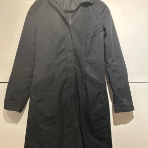 Svart lång kappa med dragkedja - Svart kappa i längre modell med snygga ribbade detaljer på både fram- och baksidan. Jackan har dragkedja framtill och är helsvart med stilren look. Materialet känns som bomull och polyester, perfekt för dig som gillar minimalistisk stil.