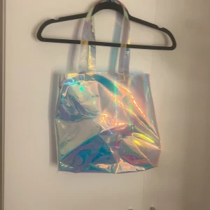 Holografisk shoppingväska i syntet - Säljer en holografisk shoppingväska med regnbågsskimrande yta. Väskan har två handtag och är tillverkad i syntetmaterial som reflekterar ljus i olika färger som rosa, blått, gult och grönt. Perfekt för dig som vill sticka ut med en unik accessoar.
