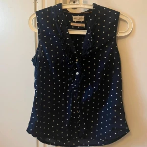 Mörkblå blus - Snygg mörkblå blus från Holly & Whyte med vita prickar, volangdetaljer längs v-ringningen och knappar framtill. Ärmlös modell i lätt och luftig bomull, perfekt för en fräsch och stilren look.