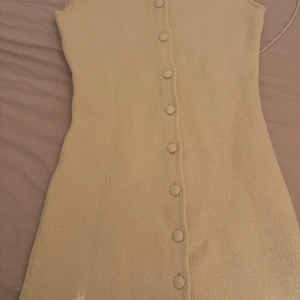 Beige ärmlös klänning med knappar - Supersöt beige klänning med ärmlös design och rund hals. Klänningen har en rak, kort siluett och är dekorerad med stora tygklädda knappar längs hela framsidan. Materialet är stickat och ger en mjuk och lite strukturerad känsla. Har aldrig använt klänningen. 