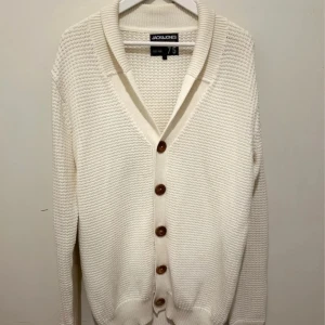 Vit kofta från Jack & Jones, storlek L - Snygg vit kofta från Jack & Jones i storlek L. Koftan är stickad i bomull och har bruna knappar framtill samt ribbade muddar. V-ringad med lång ärm och coola armbågslappar i vitt på baksidan. Perfekt för dig som gillar stilrena och klassiska plagg.