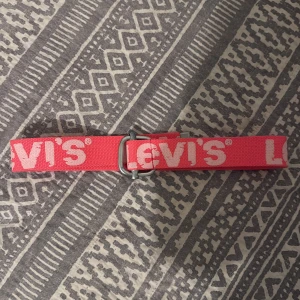 Rosa Levi's bälte med vit logga - Säljer ett snyggt rosa bälte från Levi's med stor vit logga tryckt längs hela bandet. Bältet har en metallspänne i silverfärg och är gjort i slitstark polyester. Perfekt för att ge outfiten en cool streetwear-vibe.  Bredd: 86cm. Höjd: 4cm