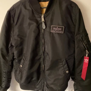 Svart bomberjacka Alpha Industries - Säljer en svart bomberjacka från Alpha Industries med klassisk patch på bröstet och röd tag på ärmen. Jackan har ribbade muddar, dragkedja framtill och fickor med tryckknappar. Perfekt för dig som gillar streetwear och snygg, enkel stil.