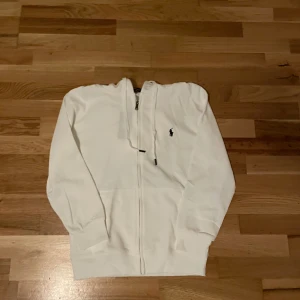 Vit hoodie från Polo Ralph Lauren - Vit hoodie från Polo Ralph Lauren med dragkedja och huva med snörning. Klassisk logga broderad på bröstet. Tröjan har långa ärmar och ribbade muddar. Tillverkad i mjuk bomull som är skön att ha på sig. Perfekt för chill dagar eller när du vill ha en clean look.
