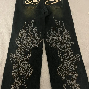 Ed Hardy jeans med drakmotiv - Säljer ett par svarta Ed Hardy jeans med stora vita drakmotiv längs benen och tydliga loggor på bakfickorna och framtill. Jeansen har en loose passform och raka ben, tillverkade i klassisk denim. Grym streetstil med unika detaljer. Endast använda få gånger. Nypris: 11059 kr. Köpta på Urban outfitters