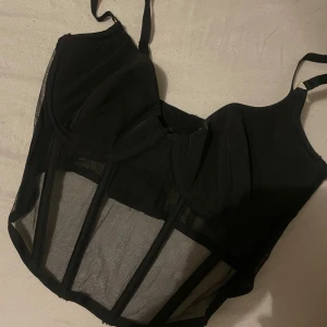 Svart mesh korsett från H&M - Snygg svart korsett från H&M med transparent mesh och markerade sömmar. Axelbanden är justerbara och kuporna har lätt vaddering för extra stöd. Perfekt för att ge en edgy vibe till din outfit.