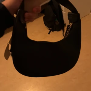 Svart axelväska med gulddetaljer - Snygg svart axelväska i skinnimitation med guldfärgade metalldetaljer och dragkedja upptill. Väskan har en stilren, rundad form och är perfekt för att bära det viktigaste. Enkel design som funkar till många olika outfits.