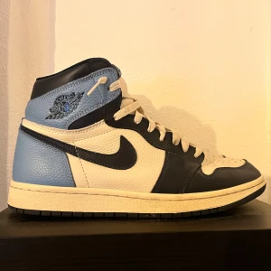 Nike Air Jordan 1 High blå/vit - Nike Air Jordan 1 High sneakers i blått, vitt och svart skinn. Klassisk siluett med högt skaft, perforerad tå och ikonisk Swoosh på sidan. Medföljer extra vita skosnören. Perfekt för dig som gillar streetwear och snygga detaljer.