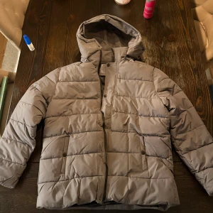 Grå pufferjacka med huva - Säljer en grå pufferjacka med huva och horisontella quiltade paneler. Jackan har dragkedja och tryckknappar framtill samt två fickor med knappstängning. Perfekt för kalla dagar och har en clean, modern look.