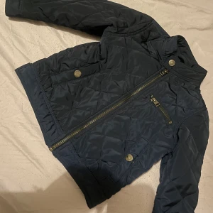 Mörkblå quiltad jacka Holly & Whyte - Quiltad mörkblå jacka från Holly & Whyte by Lindex i storlek 92. Jackan har snygga detaljer med dragkedja och tryckknappar, samt en ståkrage. Materialet är polyester och jackan har en cool, lite sportig look som funkar till många stilar. Till barn 