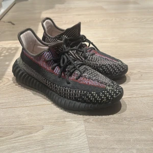 Adidas Yeezy Boost 350 V2 sneakers - Säljer ett par Adidas Yeezy Boost 350 V2 med stickad ovandel i svart, röd och beige med coolt mönster. Skorna har snörning, platt sula och den klassiska Boost-dämpningen. Perfekt för dig som gillar streetwear och vill sticka ut med en unik färgkombo. Priset kan diskuteras ganska mycket och är billigare vid smidig affär 