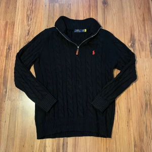 Svart stickad tröja Polo Ralph Lauren - Svart kabelstickad tröja från Polo Ralph Lauren med röd broderad logga på bröstet. Tröjan har en hög krage med half zip-dragkedja och ribbade muddar vid ärmslut och nederkant. Perfekt för dig som gillar klassisk och stilren look. Size L/180 (längd (70cm), bredd (48 cm), ärmlängd (62cm)  Pris går att diskutera 🫡