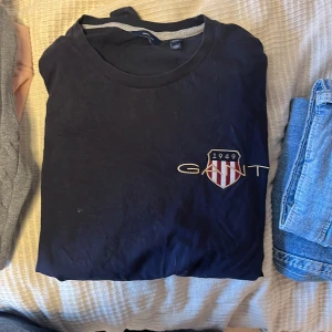 Mörkblå långärmad t-shirt från GANT  - Snygg mörkblå tröja från GANT med broderad logga och sköld på bröstet. Klassisk rund hals och mjukt bomullsmaterial som är skönt mot huden. Perfekt för dig som gillar stilrena märkesplagg med cool detalj framtill. 