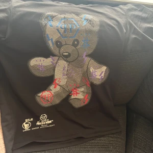 Svart Philipp Plein t-shirt med nalletryck - Cool svart t-shirt från Philipp Plein Platinumcut med stort glittrigt nalletryck framtill och färgglada blixtar i blått, rött och lila. Limited edition med exklusiva detaljer och logotyptryck. Skön bomullskvalitet och klassisk passform. Ny pris ligger på runt 5500