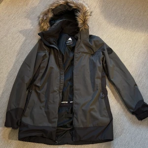 Burton - grå skidjacka  - Säljer en grå skidjacka från Burton med snygg fuskpäls på huvan och coolt logomärke på ärmen. Jackan har dragkedja, flera fickor och justerbara muddar. Perfekt för kalla dagar med sin långa passform och slitstarka material.