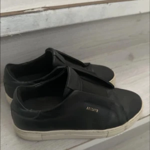Svarta sneakers från Axel Arigato - Stilrena svarta sneakers från Axel Arigato med vit sula och diskret guldfärgad logga på sidan. Skorna är i skinn och har en minimalistisk design utan snörning, vilket ger en clean och modern look. har lite skador skriv för flera bilder