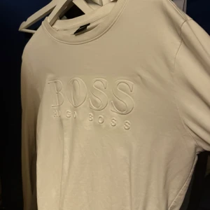 Vit långärmad tröja från Hugo Boss - Snygg vit långärmad tröja från Hugo Boss med upphöjt BOSS-tryck på bröstet. Tröjan har en klassisk rund hals och är gjord i mjuk bomull som känns skön mot huden. Perfekt för dig som gillar stilrena och enkla designer.