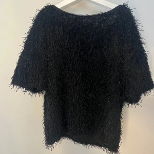 Svart fluffig topp med kort ärm - Säljer en svart fluffig topp med korta ärmar och rund hals. Toppen har en cool textur med långa, mjuka trådar som ger ett unikt och trendigt uttryck. Passformen är normal och toppen är croppad i modellen. Perfekt för dig som vill sticka ut med en annorlunda look.
